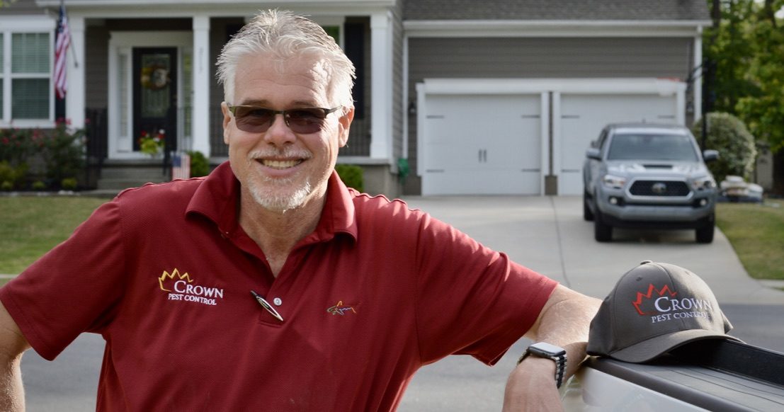 George Bryce A man wearing sunglasses and a red polo shirt with a "Crown Pest Control" logo smiles while leaning on a vehicle. There's a gray house and a pickup truck in the background. A "Crown Pest Control" hat is placed on the hood of the vehicle. George Bryce from Matthews NC proudly showcases his work attire.