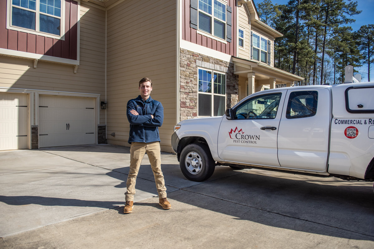 A person standing on a driveway in front of a beige house, arms crossed. Nearby is a white pickup truck with "Crown Pest Control" on the side. The house has several windows and is surrounded by tall trees.